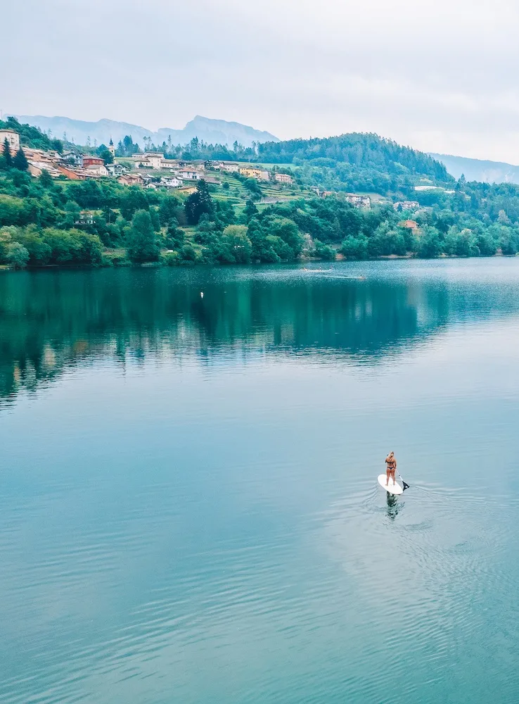 Drone shot of a girl doing SUP in the middle of Lake Caldonazzo in Trentino, Italy, with the mountains in the background and shoreline reflected in the lake