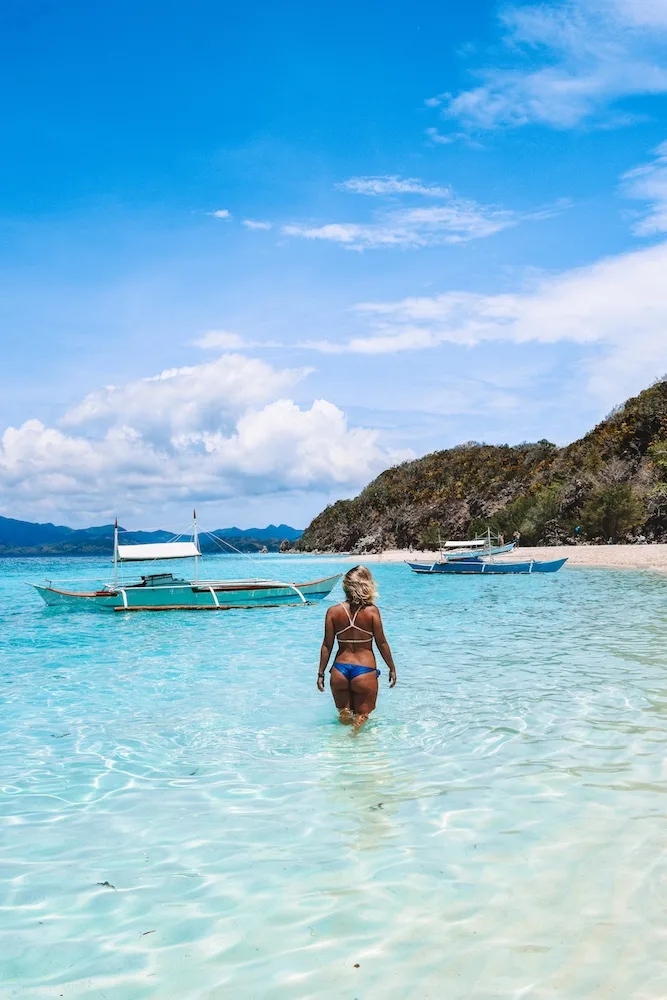 Girl walking in the clear turquoise water of Malcapuya Island, Philippines, with a typical Filipino boat behind her