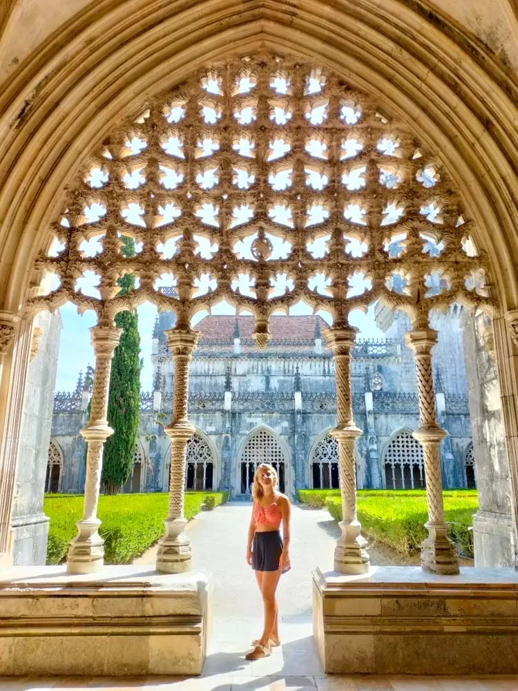 Blonde girl in a pink top and black shorts standing beneath the gothic decorations of the cloisters of Batalha monastery - a must-see on any Portugal 2-week itinerary!
